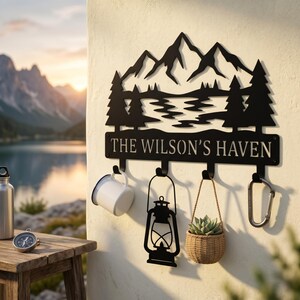 May include: Black metal wall-mounted key holder with a mountain and lake scene, the text "THE WILSON'S HAVEN", and four hooks. A white mug, a lantern, a small woven basket with a plant, and a carabiner are hanging from the hooks.