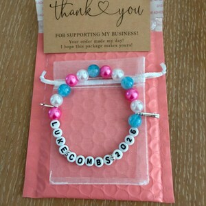 May include: A colourful beaded bracelet spelling "LUKE COMBS 2026" with white letter beads. The bracelet has pink, blue, and white beads. It's inside a small, clear bag, on a pink bubble mailer. A thank you card is on top, with a message.