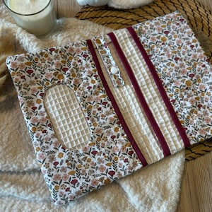 Peut inclure: Une housse de boîte à mouchoirs rectangulaire en tissu avec un motif floral composé de fleurs roses, rouges et jaunes sur fond blanc. Elle présente une ouverture ovale centrale, un panneau texturé gaufré crème et trois bandes verticales rouge foncé.