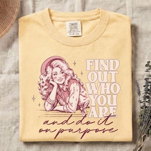 May include: A pale yellow t-shirt featuring a graphic illustration of a woman with long wavy hair wearing a hat. The text reads "FIND OUT WHO YOU ARE and do it on purpose" in a mix of block and script fonts. The shirt has a Comfort Colors label.