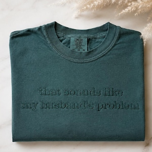 May include: A dark teal crew neck t-shirt featuring the embroidered text that sounds like my husband's problem in a matching dark thread. The shirt has a Comfort Colors label visible at the inner neckline, indicating a medium size.