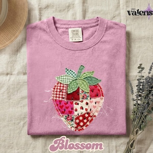 May include: A light pink t-shirt with a patchwork strawberry design. The strawberry is made of various fabric patterns, including red gingham, floral, and heart prints. The word "Blossom" is written in a cursive font.