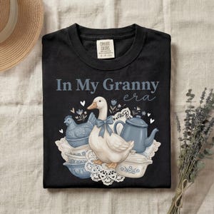 May include: Black t-shirt with the phrase "In My Granny era" and a graphic of a goose with a blue bow, a blue chicken, and blue teacups. The design includes white lace and blue floral accents.