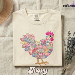 May include: Ivory-colored t-shirt featuring a colorful floral hen design. The hen is made of various fabric patterns in pink, blue, and yellow. The shirt has a Comfort Colors tag and the word "Ivory" at the bottom.