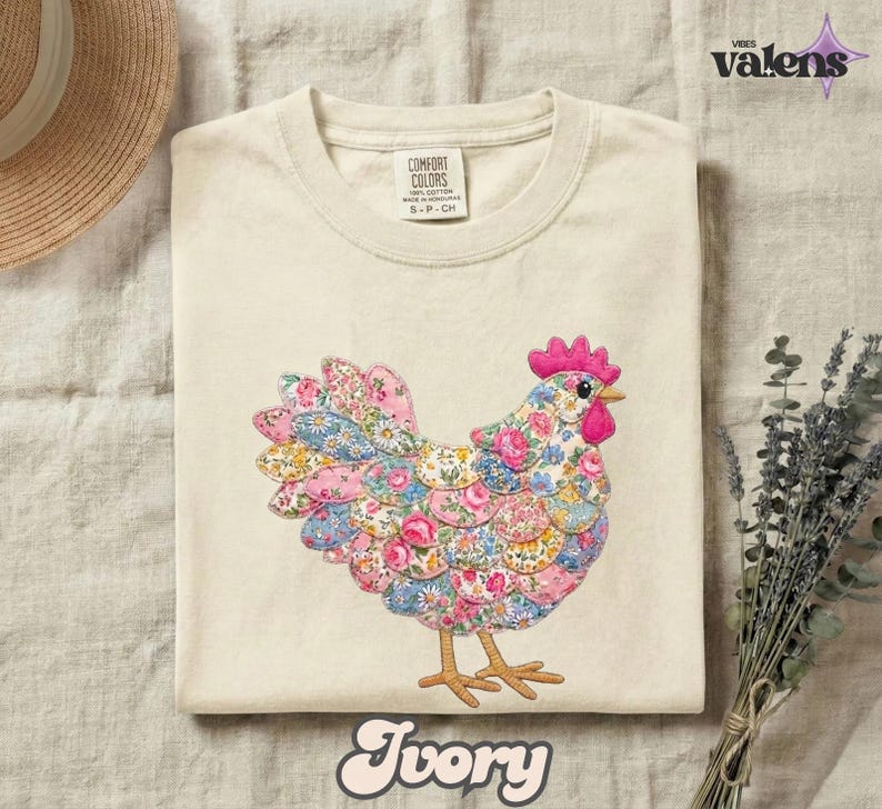 May include: Ivory-colored t-shirt featuring a colorful floral hen design. The hen is made of various fabric patterns in pink, blue, and yellow. The shirt has a Comfort Colors tag and the word "Ivory" at the bottom.