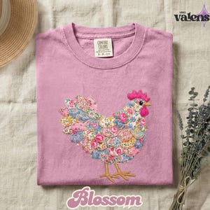 May include: A pink Comfort Colors t-shirt featuring an embroidered hen with a floral pattern in shades of pink, blue, and yellow. The hen has a red comb and beak. The word "Blossom" is written in a cursive font at the bottom of the shirt.