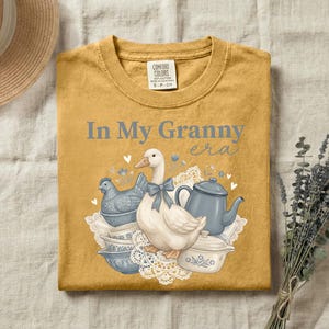 May include: Mustard yellow t-shirt with the phrase "In My Granny era" in light blue. The graphic features a goose with a blue bow, a blue chicken-shaped dish, and blue and white teacups and teapot. The shirt is a Comfort Colors brand.