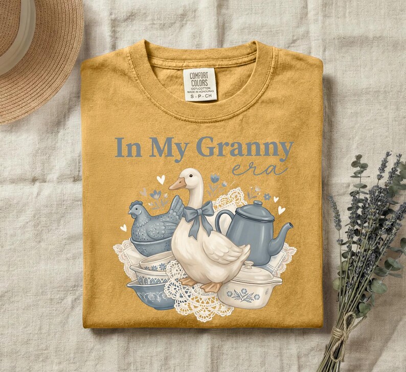 May include: Mustard yellow t-shirt with the phrase "In My Granny era" in light blue. The graphic features a goose with a blue bow, a blue chicken-shaped dish, and blue and white teacups and teapot. The shirt is a Comfort Colors brand.
