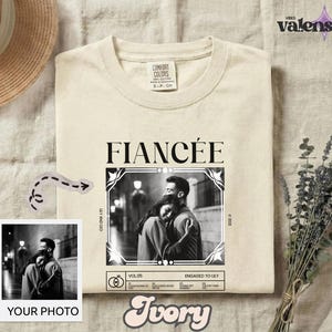 May include: A cream-colored t-shirt featuring a black graphic print with the word FIANCÉE in a serif font above a framed black and white photograph of a couple embracing. The design includes text reading LILY AND LEO, VOL.05, and ENGAGED TO LILY.