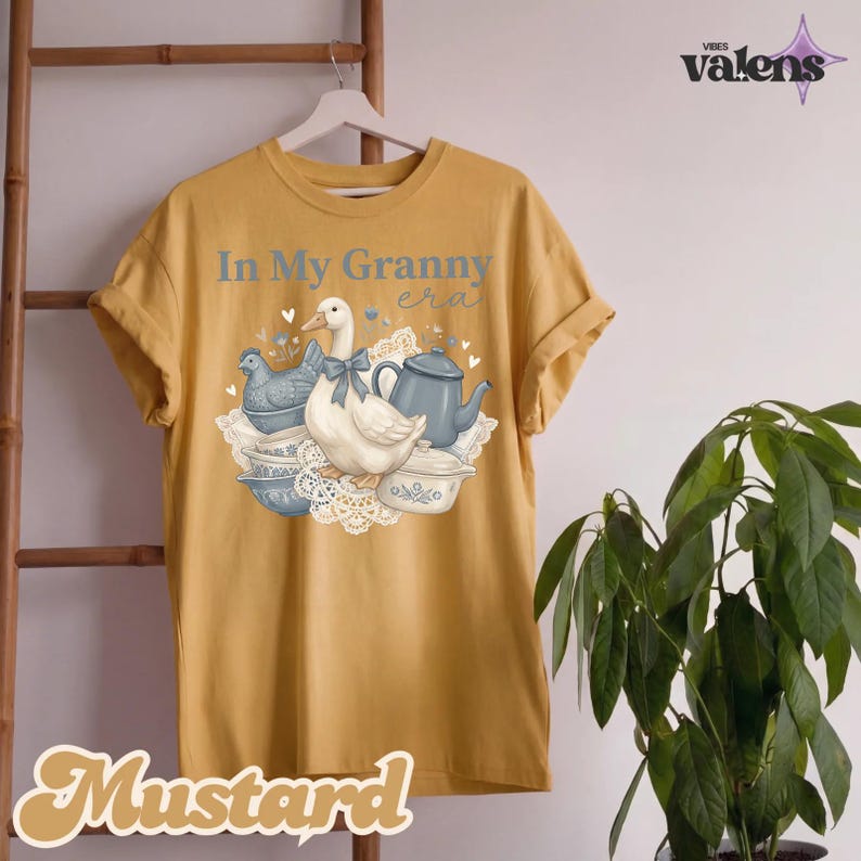 May include: Mustard-colored t-shirt with rolled-up sleeves. The front features a graphic of a white duck with a blue bow, a teapot, and the text "In My Granny era". The word "Mustard" is written in a decorative font at the bottom.