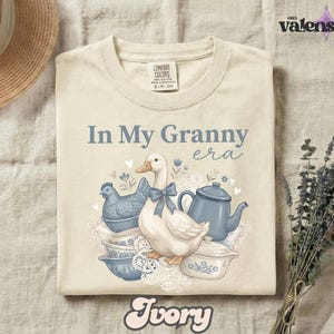 May include: Ivory-colored t-shirt with the phrase "In My Granny era" in blue script. The graphic features a white duck with a blue bow, blue teacups, and a teapot. The shirt is a Comfort Colors brand.