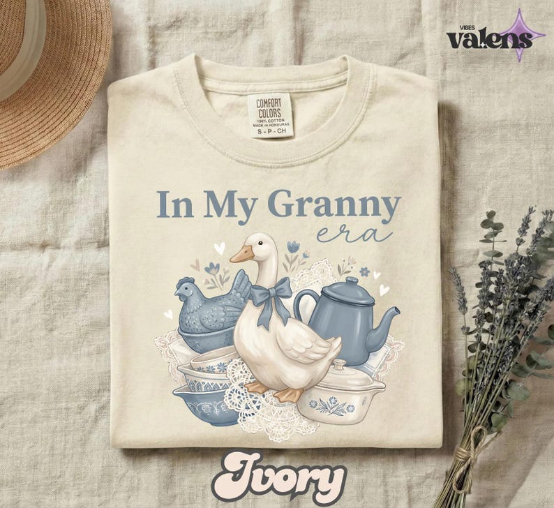 May include: Ivory-colored t-shirt with the phrase "In My Granny era" in blue script. The graphic features a white duck with a blue bow, blue teacups, and a teapot. The shirt is a Comfort Colors brand.