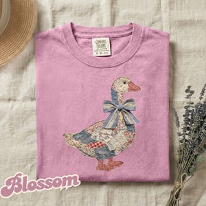 May include: A pink Comfort Colors t-shirt featuring an appliqued goose design. The goose is made of patchwork fabric with a blue gingham bow. The word "Blossom" is written in a cursive font at the bottom left.