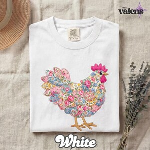May include: White t-shirt featuring a colorful floral chicken design. The chicken has a pink comb and wattle, with a body made of various floral patterns in pink, blue, and yellow. The shirt has a Comfort Colors tag and the word "White" at the bottom.