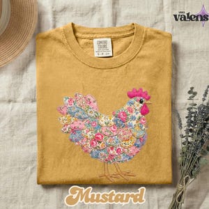 May include: Mustard-colored t-shirt with a floral chicken design. The chicken is made of various floral patterns in pink, blue, and white. The shirt has a crew neck and short sleeves. The text "Mustard" is at the bottom.