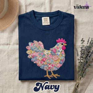 May include: Navy blue t-shirt featuring a colorful floral chicken applique. The chicken design incorporates pink, blue, and yellow floral patterns. The shirt has a crew neck and short sleeves. The word "Navy" is written in a cursive font at the bottom.