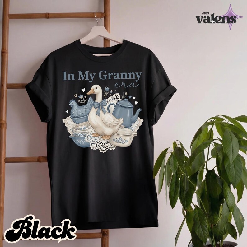 May include: Black t-shirt with the text "In My Granny era" and a graphic of a goose with a blue bow, a teapot, and a chicken. The graphic is in shades of blue and white. The word "Black" is written in a cursive font at the bottom.