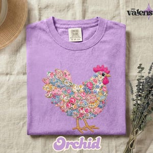 May include: A lilac-colored Comfort Colors t-shirt featuring a colorful floral chicken design. The chicken has a pink comb and beak, with a body made of various floral patterns. The word "Orchid" is written in a cursive font at the bottom.