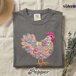 May include: A gray Comfort Colors t-shirt featuring a colorful floral chicken design. The chicken has a pink comb and wattle, with a body made of pink, blue, and yellow floral fabric. The word "Pepper" is written in a script font at the bottom.