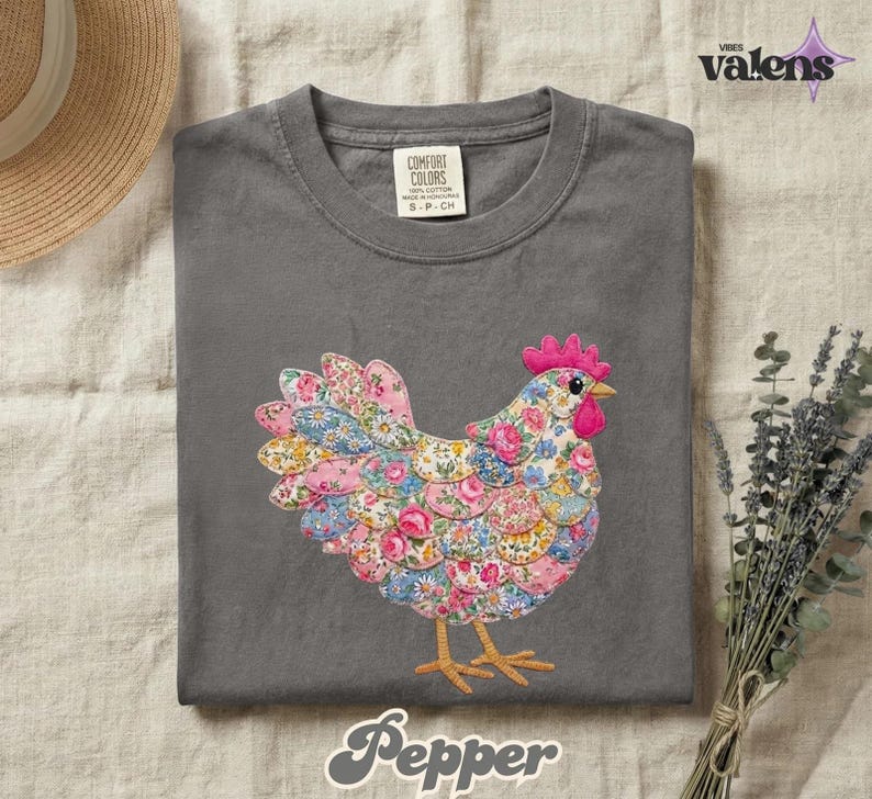 May include: A gray Comfort Colors t-shirt featuring a colorful floral chicken design. The chicken has a pink comb and wattle, with a body made of pink, blue, and yellow floral fabric. The word "Pepper" is written in a script font at the bottom.