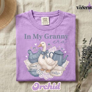 May include: A lilac-colored t-shirt with the phrase "In My Granny Era" and a graphic of a goose, teapot, and other vintage kitchen items. The word "Orchid" is at the bottom of the shirt.