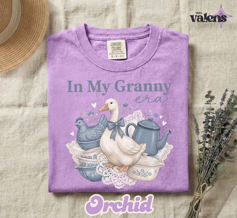 May include: A lilac-colored t-shirt with the phrase "In My Granny Era" and a graphic of a goose, teapot, and other vintage kitchen items. The word "Orchid" is at the bottom of the shirt.