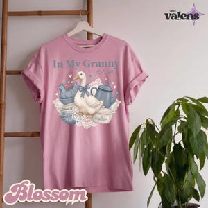 May include: A pink t-shirt with the text "In My Granny era" and a graphic of a duck, chicken, teapot, and bowls. The design features a duck with a blue bow, a blue chicken, and a blue teapot. The word "Blossom" is at the bottom.