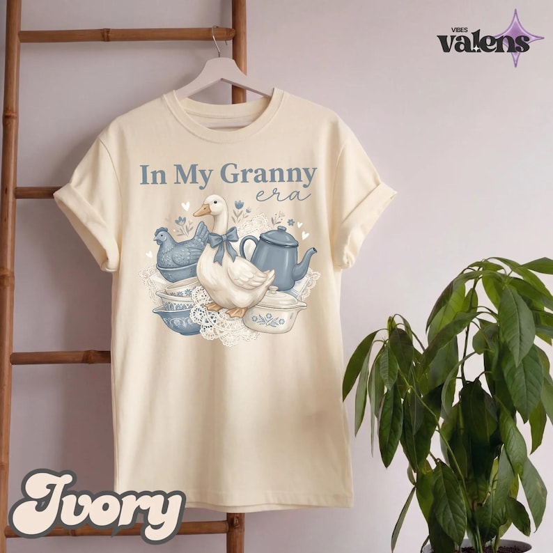 May include: Ivory-colored t-shirt with the text "In My Granny era" and a graphic of a duck, teapot, and dishes. The graphic is in shades of blue and white. The shirt is hanging on a wooden hanger.