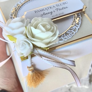 Könnte beinhalten: Eine quadratische, cremefarbene Geschenkbox mit drei weißen Kunstrosen, einem weiß-goldenen Band und einer kleinen goldenen Quaste. Auf der Oberseite befindet sich ein weißes Etikett mit dem Text PAMIĄTKA ŚLUBU Anny i Piotra 20.06.2026.
