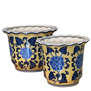 May include: Two yellow ceramic flower pots with blue floral designs. The pots have a scalloped rim and a white interior. The blue floral pattern includes large flowers and leafy vines. The pots are likely for indoor or outdoor use.