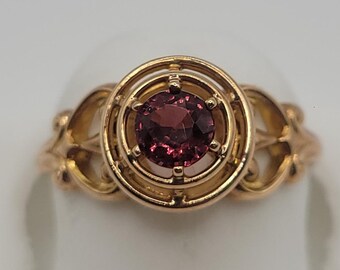Puede incluir: Un anillo de oro con una piedra preciosa redonda de color rojo oscuro engastada en un marco circular. La banda presenta un diseño calado intrincado con elementos metálicos curvos que se extienden desde el engaste central, creando una apariencia detallada.