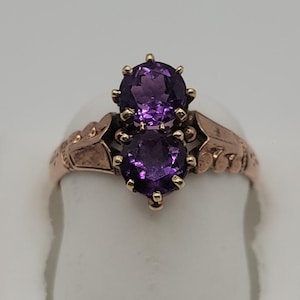 May include: A gold ring featuring two round, faceted purple amethyst gemstones set vertically in a prong setting. The metal band displays decorative, engraved leaf-like details on both sides of the central stones, creating a distinct, structured appearance.