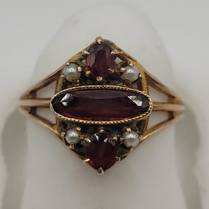 May include: A gold ring featuring a central horizontal oval-cut deep red garnet, flanked by two smaller vertical oval-cut garnets at the top and bottom. Four small white seed pearls are set between the garnets, all mounted on a split gold band.