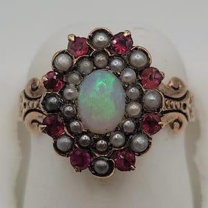 Può includere: Un anello in oro con un opale ovale centrale circondato da un alone di piccole perle bianche e un bordo esterno di gemme sfaccettate rosso scuro. La fascia in metallo presenta intricati motivi a volute incisi sui lati della montatura.