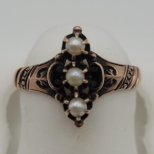 May include: A gold ring featuring an elongated, dark-toned metal setting with intricate scrollwork patterns. The center of the ring is vertically aligned with three small, round, white pearls held in place by prongs within the dark metal design.
