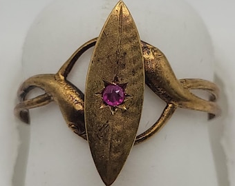 Puede incluir: Un anillo de tono dorado que presenta una placa central en forma de marquesa con una superficie texturizada. Una pequeña piedra preciosa redonda de color rosa vibrante está engastada en el centro. El aro tiene dos figuras curvas de delfines.