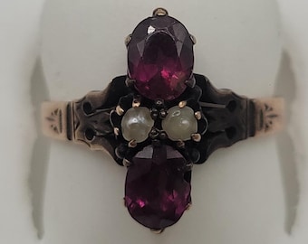 Puede incluir: Un anillo con dos piedras granate de color rojo intenso y corte ovalado dispuestas verticalmente, separadas por dos pequeñas perlas blancas redondas. El engaste es de metal oscuro con grabados decorativos en los lados del aro.