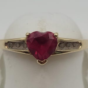 May include: A gold ring featuring a central heart-shaped red gemstone held in a prong setting. The band is crafted from yellow gold and includes a channel-set row of three small clear stones on each side of the central heart-shaped red stone.