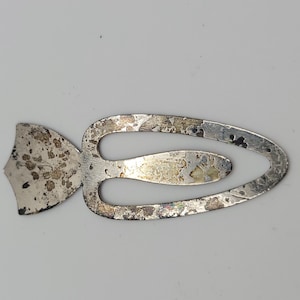 May include: A silver-toned metal bookmark featuring a flat, shield-shaped end attached to an elongated, oval-shaped loop. The surface of the metal shows visible signs of wear, including dark spots, tarnishing, and a weathered, aged texture.