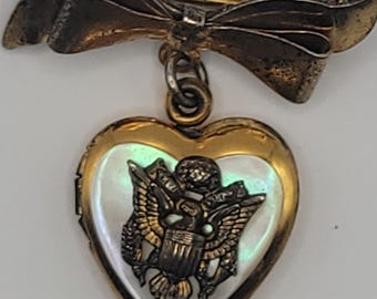 May include: A gold-toned metal brooch featuring a bow at the top from which a heart-shaped locket hangs. The locket has a white iridescent inlay and a central metallic emblem depicting an eagle with spread wings and a shield on its chest.