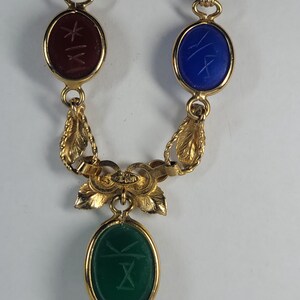 Puede incluir: Un collar de tono dorado con tres piedras ovaladas en cabuj&oacute;n de color rojo oscuro, azul y verde. Cada piedra est&aacute; grabada con un s&iacute;mbolo y engastada en un marco de metal dorado. La cadena incluye eslabones decorativos en forma de hoja.