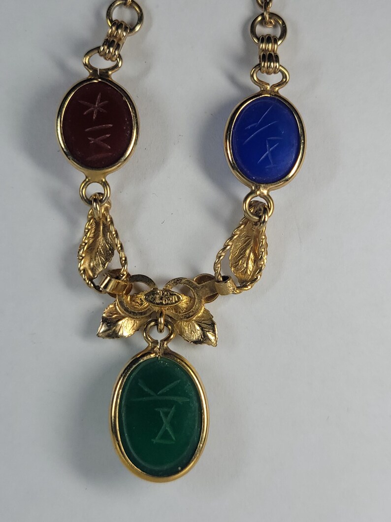 Puede incluir: Un collar de tono dorado con tres piedras ovaladas en cabuj&oacute;n de color rojo oscuro, azul y verde. Cada piedra est&aacute; grabada con un s&iacute;mbolo y engastada en un marco de metal dorado. La cadena incluye eslabones decorativos en forma de hoja.