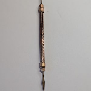 May include: A copper-toned, double-ended tool featuring a spoon-shaped end and a pointed tip. The central section has a textured pattern. This tool, likely for scooping or scraping, has a vintage design. The length is approximately 15 cm.