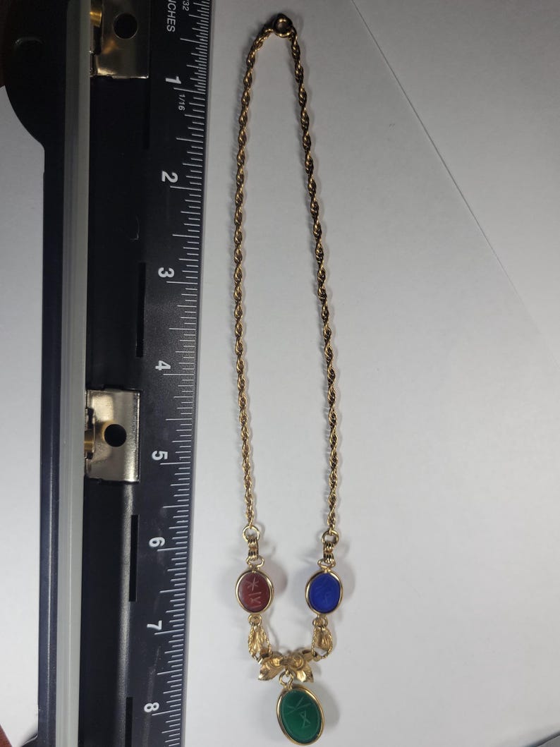 Puede incluir: Un collar de cadena trenzada dorada con tres piedras cabuj&oacute;n ovaladas en rojo, azul y verde, con un detalle met&aacute;lico floral sobre la piedra verde. Una regla negra muestra una longitud de aproximadamente 20,3 cm.