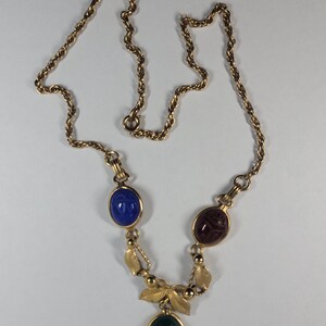 Puede incluir: Collar dorado con una cadena de cuerda trenzada. El colgante presenta tres piedras preciosas ovaladas en azul, rojo y verde. El collar tiene un dise&ntilde;o de hojas y cuentas. Las piedras est&aacute;n engastadas en metal dorado.