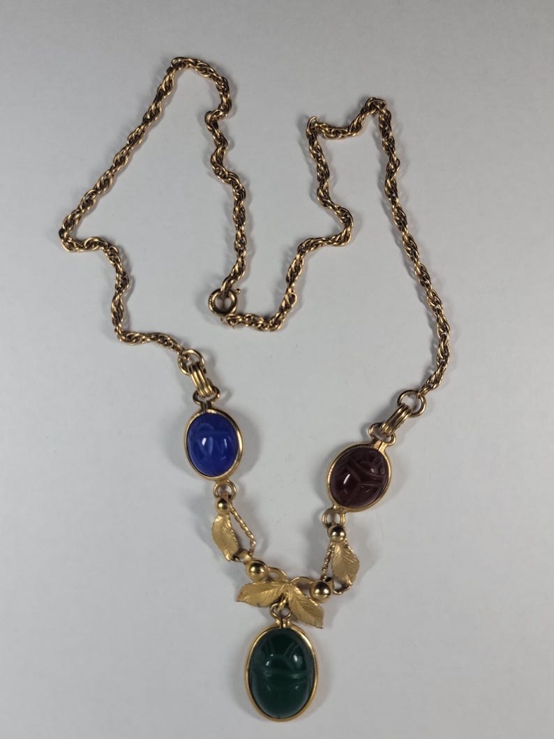 Puede incluir: Collar dorado con una cadena de cuerda trenzada. El colgante presenta tres piedras preciosas ovaladas en azul, rojo y verde. El collar tiene un dise&ntilde;o de hojas y cuentas. Las piedras est&aacute;n engastadas en metal dorado.