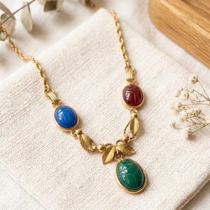 Puede incluir: Un collar de tono dorado con tres cabujones ovalados estilo escarabajo en rojo, azul y verde. La cadena est&aacute; decorada con peque&ntilde;as hojas de metal dorado y cuentas esf&eacute;ricas, conectando las piedras de colores en una disposici&oacute;n sim&eacute;trica.