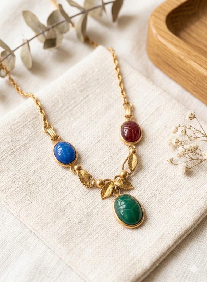Puede incluir: Un collar de tono dorado con tres cabujones ovalados estilo escarabajo en rojo, azul y verde. La cadena est&aacute; decorada con peque&ntilde;as hojas de metal dorado y cuentas esf&eacute;ricas, conectando las piedras de colores en una disposici&oacute;n sim&eacute;trica.