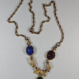 Puede incluir: Collar dorado con tres colgantes ovalados de piedras preciosas azules, burdeos y verdes. El collar tiene una cadena delicada y detalles de hojas decorativas, creando una est&eacute;tica elegante y vintage.