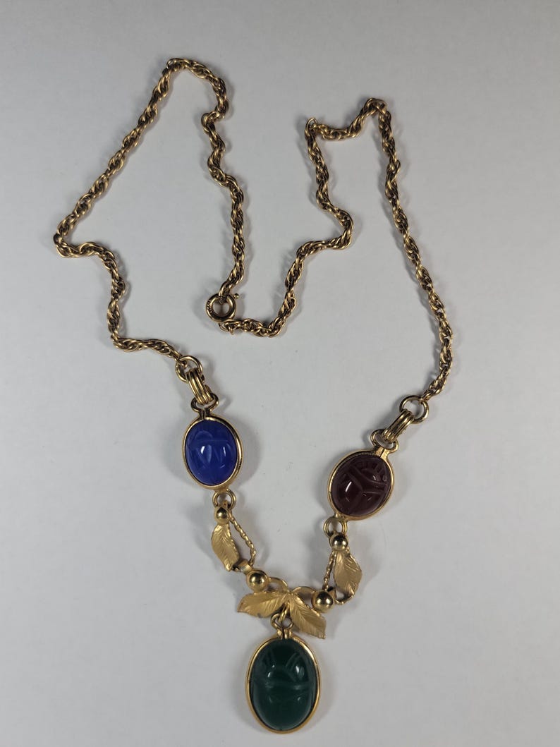 Puede incluir: Collar dorado con tres colgantes ovalados de piedras preciosas azules, burdeos y verdes. El collar tiene una cadena delicada y detalles de hojas decorativas, creando una est&eacute;tica elegante y vintage.